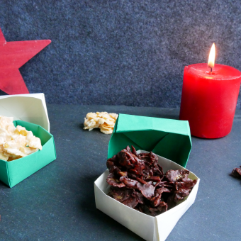 Mandel-Schoko-Crossies mit weißer und dunkler Kuvertüre in selbstgebastelter grün-weißen Schachtel. Im Hintergrund steht ein roter Stern und es brennt eine rote Kerze.