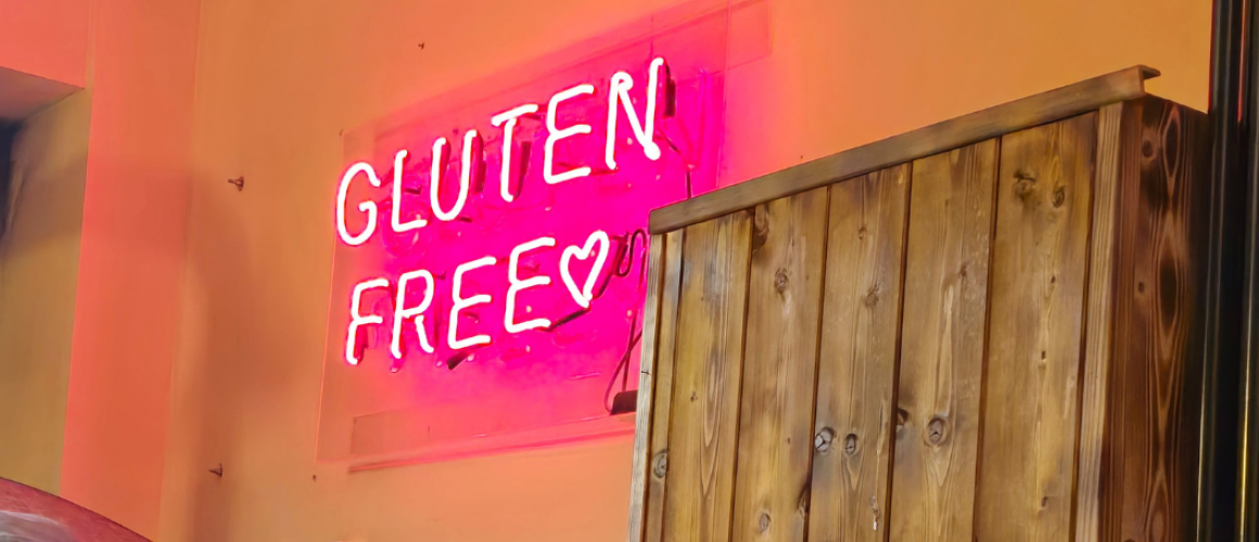 Pinkfarbene Leuchtschrift Glutenfree an einer ockerfarbenen Wand. Neben der Leuchtschrift steht ein robuster Holzschrank